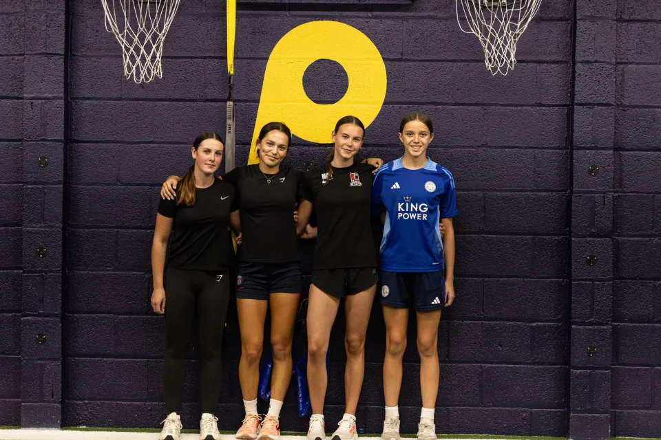 Four female athletes together beneath the Playermade logo in the gym